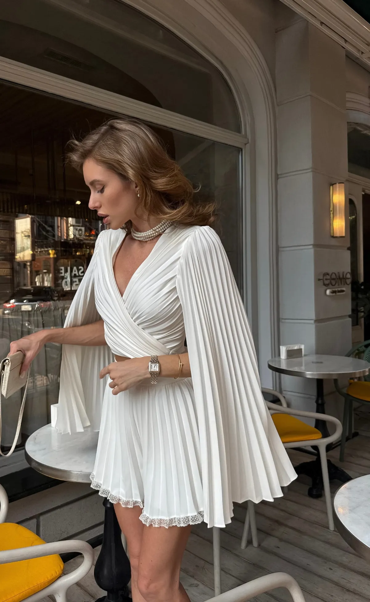 Jeune femme en ensemble plissé blanc Dream de REPULO'S avec un collier de perles sur une terrasse de café d'été